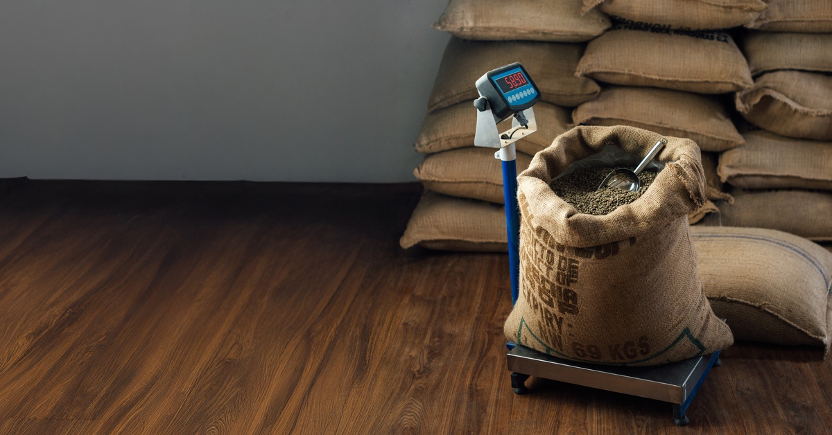 A burlap sack filled with coffee beans rests on a digital scale in a warehouse, surrounded by similar sacks and buckets.