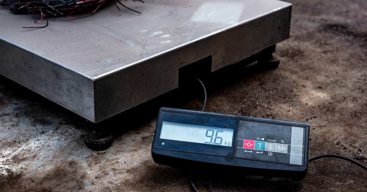 A digital platform scale displays "9.6 kg" on its screen while sitting on a concrete floor, with buttons and cables visible.