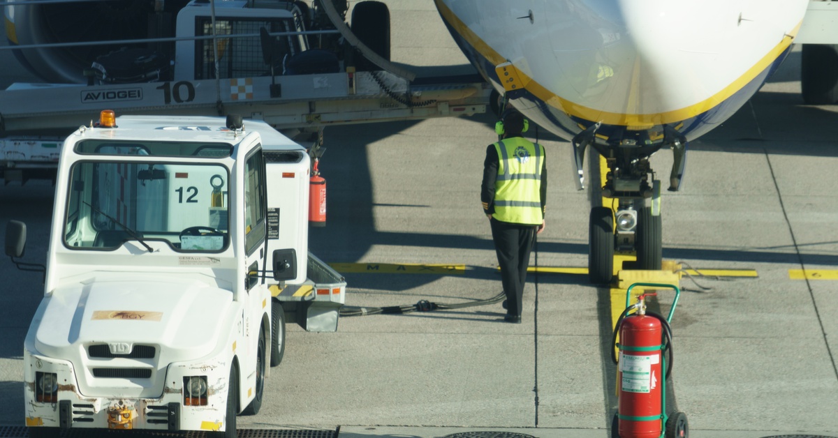 A commercial airplane parks on an airport tarmac, with a ground support vehicle and crewmembers nearby.