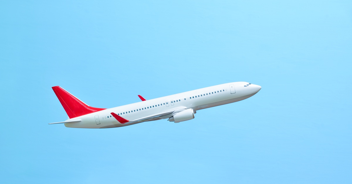 A white airplane with a red tail fin flies against a clear blue sky. The plane ascends at a diagonal angle.