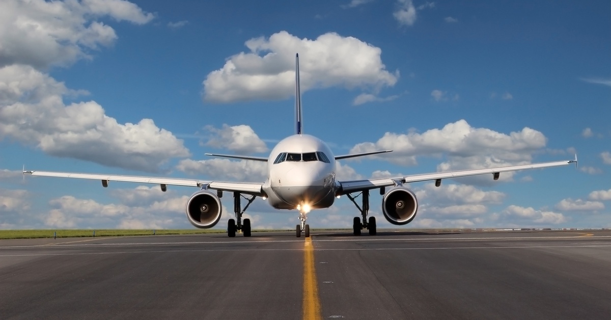 A head-on view of a white airplane with blue accents moving on the tarmac. Its landing lights illuminate under a cloudy sky.