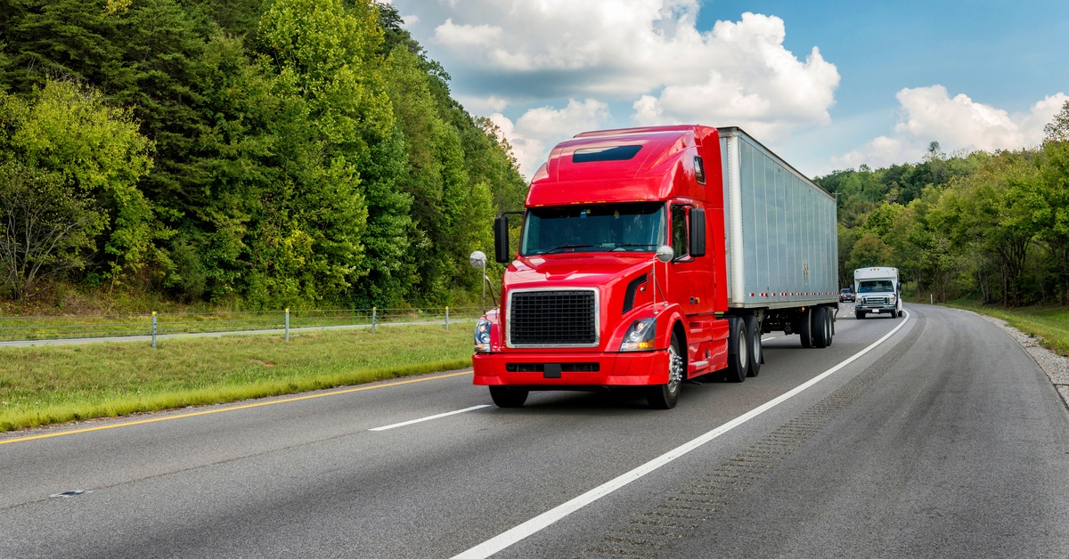 A red semi-truck with a white trailer drives on a highway surrounded by green trees and hills under a blue sky.