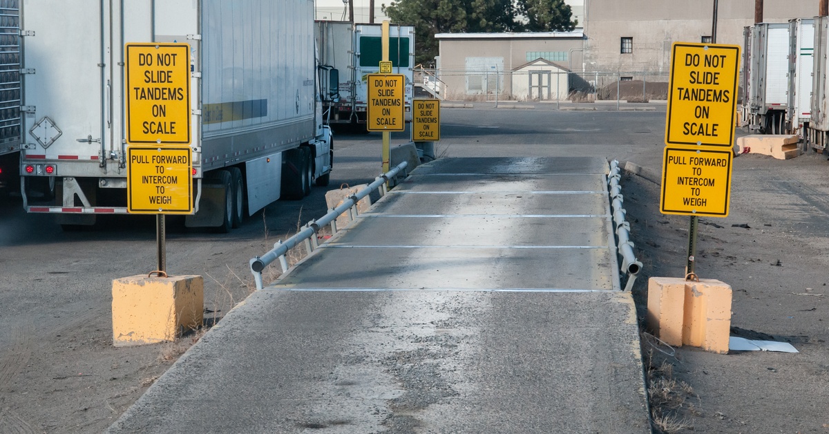 An empty truck weighing scale lane with yellow warning signs and barriers, with trailers and buildings in the background.