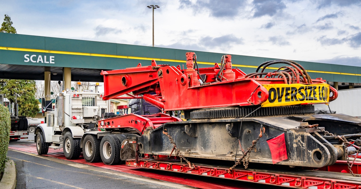 A white semi-truck pulling a trailer with large red oversized equipment secured on it; it’s parked near a weighing station.