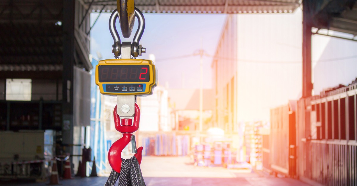 A hanging digital crane scale with a hook sits suspended in an industrial space with bright light in the background.