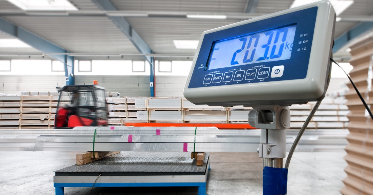A digital platform scale displays a weight reading while stacked materials sit on the scale in a warehouse.